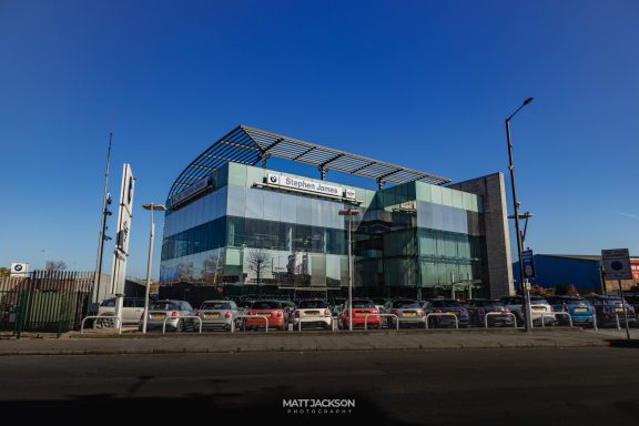 Modern glass-fronted building under a clear blue sky, with parked cars in front.